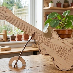 May include: A wooden guitar-shaped decorative item, resting on a wooden stand. The guitar features etched details of strings, pickups, and tuning pegs. The item is made of light-colored wood and is displayed on a wooden table.
