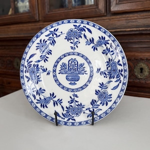 May include: White ceramic plate with a blue floral design. The plate features a central motif of a basket of flowers and is surrounded by a border of flowers and leaves. The plate is displayed on a black metal stand.