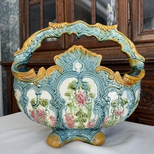 May include: An antique ceramic basket with a handle, featuring a floral design in pink, white, and green. The basket has a turquoise base and handle with gold accents. The decorative piece is likely for display.
