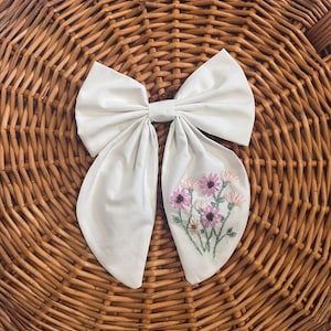 May include: A large, off-white bow with a floral embroidery of pink and white flowers. The bow is set against a woven wicker basket background, creating a contrast in textures and colors. The bow's design is simple yet elegant.