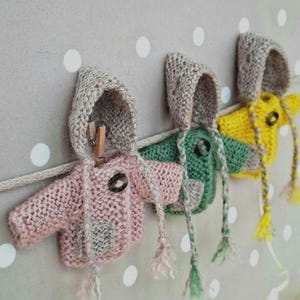 Op de afbeelding: Drie miniatuur gebreide truien met capuchon in roze, groen en geel, elk met een grijze capuchon en een knoopsluiting. De truien hangen aan een waslijn met een houten knijper. De achtergrond is lichtgrijs met witte stippen.