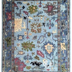 Peut inclure: Tapis rectangulaire bleu clair avec un motif floral et géométrique complexe. Le tapis présente un champ central avec diverses formes dans des tons de vert, marron et rouge, encadré par une bordure bleu foncé. Le design du tapis comprend des fleurs stylisées et d'autres éléments décoratifs.