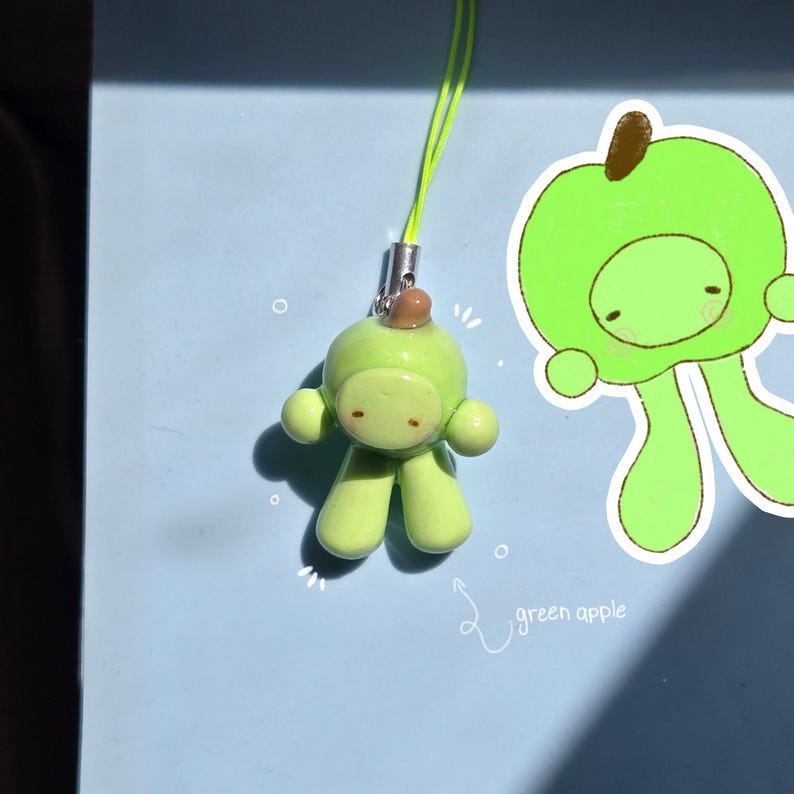May include: A light green apple-shaped charm with a small brown stem, attached to a neon yellow cord. Beside it is a white sticker featuring a matching green apple character with a brown stem and the handwritten text green apple in white.