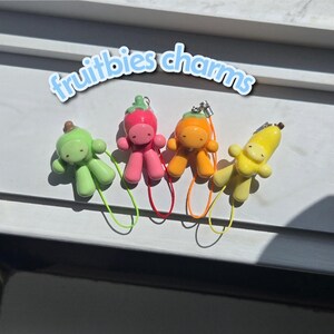 May include: Four small fruit-themed charms labeled fruitbies charms. The charms are shaped like stylized figures in light green, pink, orange, and yellow, each featuring a small stem on top and a thin colored cord loop for attachment to various items.