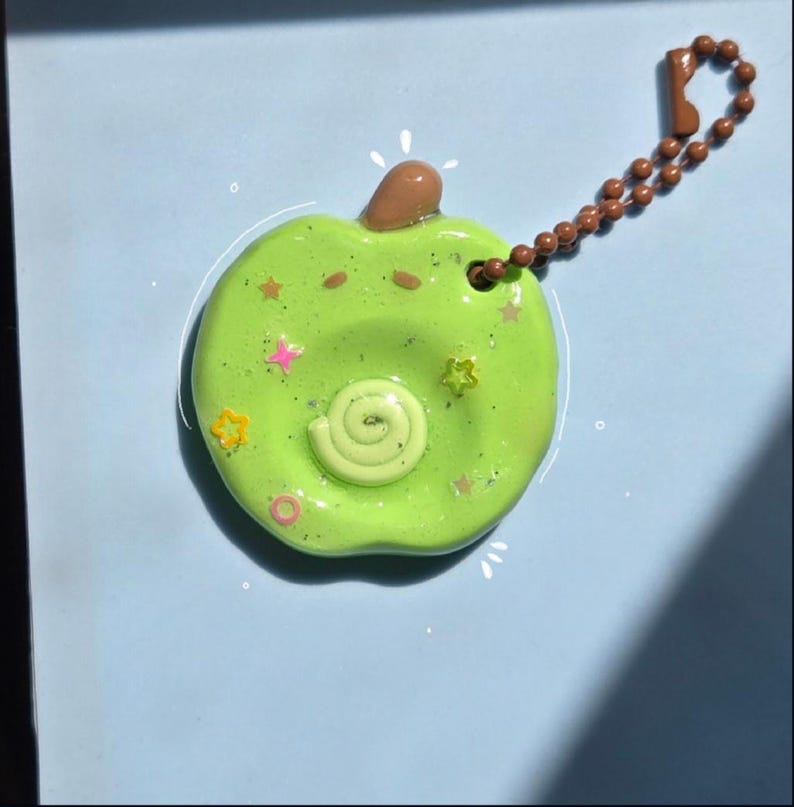 May include: A lime green apple-shaped keychain with a brown stem and a brown ball chain. The surface features a central spiral, small glittery star-shaped accents in yellow and pink, and two small brown dots representing eyes on a glossy finish.