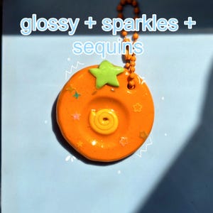 May include: A round orange keychain with a glossy finish, featuring a light green star at the top and a yellow spiral in the center. The surface is embedded with small colorful star-shaped sequins. The text glossy + sparkles + sequins is visible.