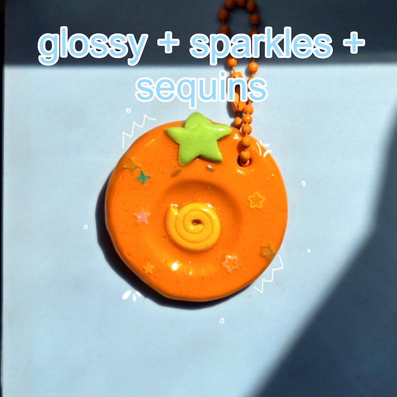 May include: A round orange keychain with a glossy finish, featuring a light green star at the top and a yellow spiral in the center. The surface is embedded with small colorful star-shaped sequins. The text glossy + sparkles + sequins is visible.