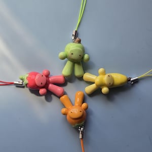 May include: Four small, smooth, clay-like charms shaped like stylized figures, each attached to a thin cord. The figures are colored light green, bright pink, yellow, and orange, featuring simple facial features and rounded limbs.