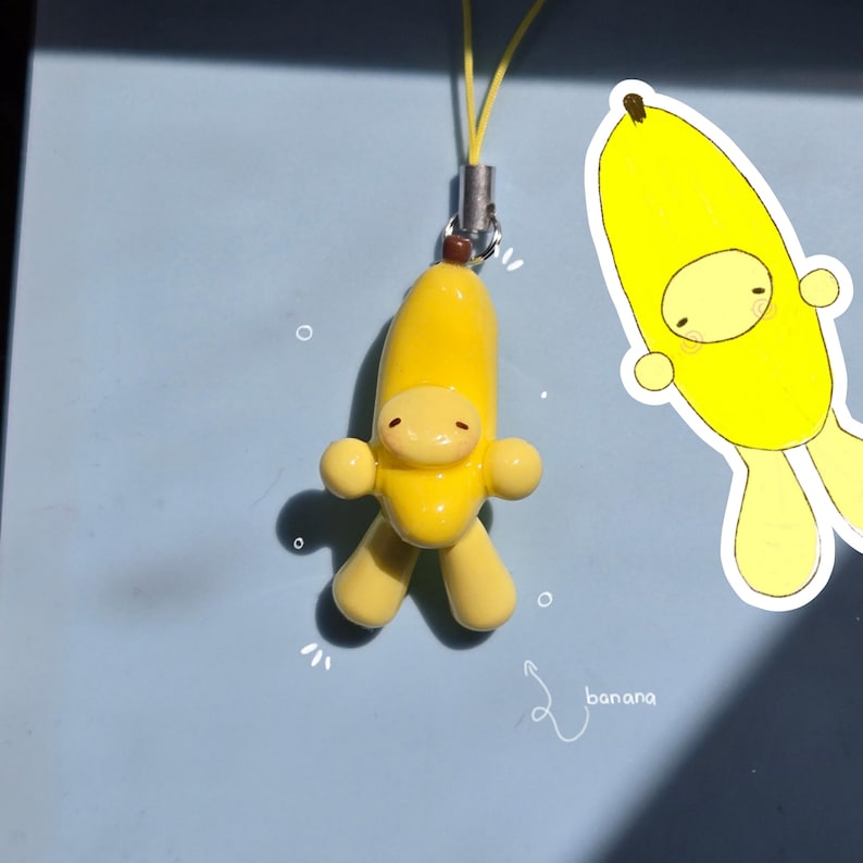 May include: A yellow banana-shaped charm featuring a small character with a smooth finish, attached to a thin yellow cord. Next to the charm is a white-bordered illustration of the same banana character with the handwritten text banana below it.