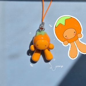 May include: A small orange fruit-shaped charm with a green top and a smooth finish, attached to a thin orange cord. Next to the charm is a hand-drawn illustration of the same character with the word orange written in white cursive script.