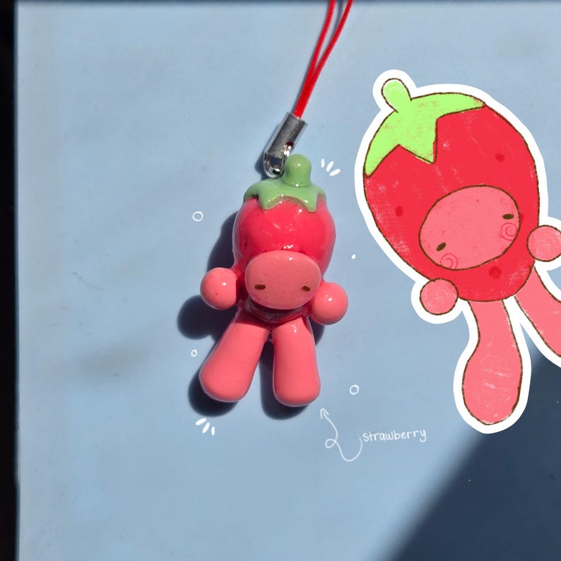 May include: A small, glossy, pink figurine shaped like a character wearing a red strawberry hat with a green stem, attached to a red string loop. Next to it is a matching hand-drawn illustration of the same character with the word strawberry.
