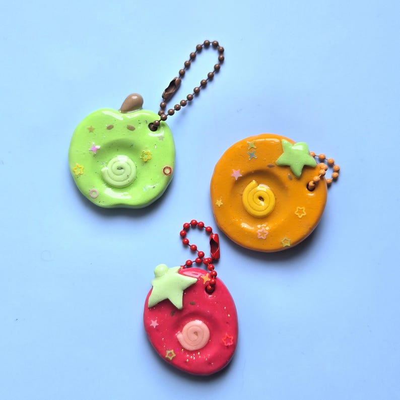 May include: Three fruit-shaped keychains featuring a lime green apple, an orange citrus slice, and a red strawberry. Each charm has a central spiral, small star-shaped glitter accents, and a metal ball chain for attachment to keys or bags.