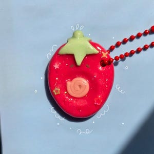 May include: A bright red strawberry-shaped pendant on a red ball chain necklace. The pendant features a light green stem, a small peach-colored spiral detail in the center, and embedded glittery star-shaped accents. White hand-drawn doodle lines surround the pendant.
