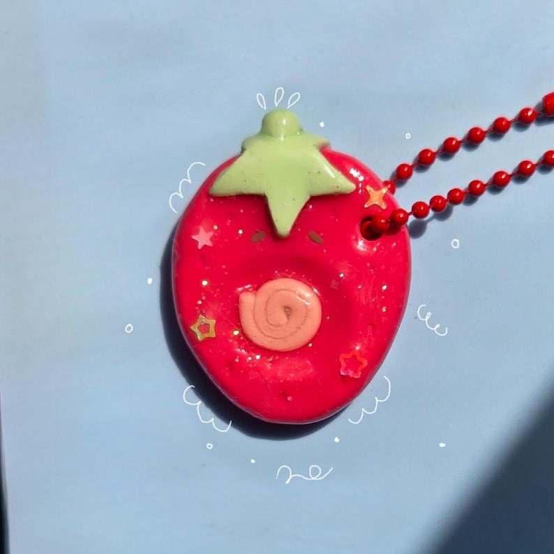 May include: A bright red strawberry-shaped pendant on a red ball chain necklace. The pendant features a light green stem, a small peach-colored spiral detail in the center, and embedded glittery star-shaped accents. White hand-drawn doodle lines surround the pendant.
