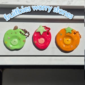 May include: Three fruit-shaped worry stones with the text "fruitbies worry stones" above them. The green apple, red strawberry, and orange citrus shapes feature a central indentation with a swirl, small star-shaped glitter, and a ball chain.