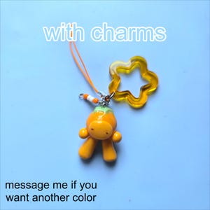 May include: An orange character charm with a green top, attached to an orange string loop and a translucent yellow star-shaped charm. The text "with charms" appears at the top and "message me if you want another color" at the bottom.