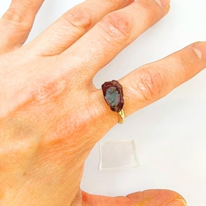 以下が含まれることがあります： 金色のリング。形が不規則な、濃い赤茶色の原石に青緑色のインクルージョンが入っています。白い背景の中で指にはめられています。原石の長さは約2cmです。
