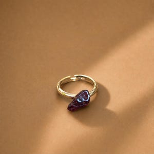 以下が含まれることがあります： 暗い紫色の、不規則な形の石が付いたゴールドカラーのリング。リングは暖かい茶色の背景に配置され、微妙な影が深みを加えています。石はユニークで有機的な形をしています。