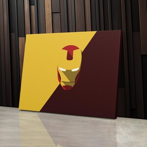 May include: A canvas art print featuring a stylized Iron Man helmet design. The artwork is divided diagonally, with a yellow section and a maroon section. The helmet is red and gold, with white highlights. The artwork is displayed on a white surface.