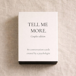 Könnte beinhalten: Eine weiße Box mit dem Text "TELL ME MORE. Couples edition" in Schwarz. Die Box enthält auch den Text "80 Gesprächskarten erstellt von einem Psychologen". Die Box liegt auf einer neutralfarbenen Oberfläche.