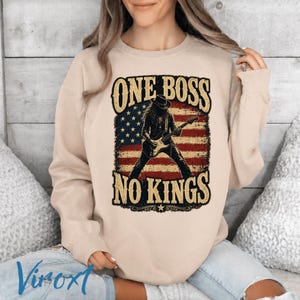 Może przedstawiać: Beżowa bluza z nadrukiem „ONE BOSS NO KINGS”. Grafika przedstawia osobę grającą na gitarze na tle amerykańskiej flagi. Bluza ma swobodny styl.