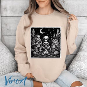 May include: Beige sweatshirt featuring a black and white graphic of a Bigfoot, an alien, and an owl knitting around a campfire under a starry night sky. The design includes a crescent moon and pine trees.