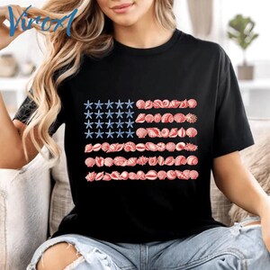 May include: A black short-sleeved t-shirt featuring a graphic design of the American flag composed of blue starfish in the upper left corner and rows of red and white seashells forming the stripes across the center of the chest area.