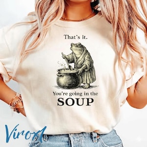 Puede incluir: Camiseta beige con un gráfico humorístico de una rana con una bata removiendo una olla. El texto de arriba dice "That's it." y debajo "You're going in the SOUP."