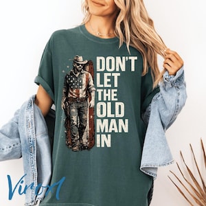 Può includere: T-shirt verde scuro con la scritta bianca "DON'T LET THE OLD MAN IN". La maglietta presenta una grafica di un uomo con cappello da cowboy e occhiali da sole, con un disegno della bandiera americana. La maglietta è uno stile casual e confortevole.