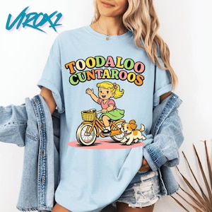May include: Light blue t-shirt featuring a cartoon illustration of a girl riding a bicycle with a dog. The text "TOODALOO CUNTAROOS" is above the illustration in colorful letters. The shirt has short sleeves and a relaxed fit.