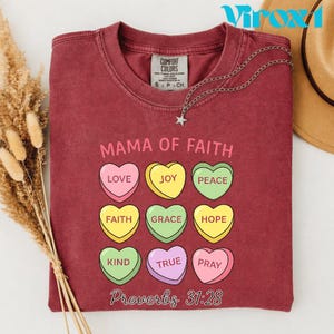 Puede incluir: Una camiseta Comfort Colors granate con las palabras "MAMA OF FAITH" y el versículo bíblico Proverbios 31:28 impresos. La camiseta presenta caramelos en forma de corazón con palabras como "LOVE", "JOY" y "PEACE". Un collar plateado con un colgante de estrella está sobre la camiseta.