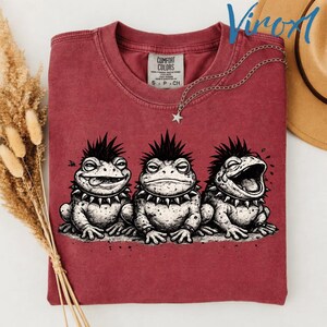 May include: A maroon t-shirt featuring a graphic of three punk rock frogs with spiky mohawks and studded collars. The shirt has a Comfort Colors tag and a silver necklace with a star pendant.
