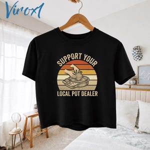May include: A black short-sleeve t-shirt featuring a graphic of hands shaping clay on a pottery wheel against a sunset-colored circle. The text reads SUPPORT YOUR LOCAL POT DEALER in a bold, arched font above and below the central illustration.