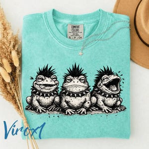 May include: A turquoise Comfort Colors t-shirt featuring a graphic of three punk rock frogs with spiky collars and mohawk hairstyles. The shirt has a small Comfort Colors tag and a silver necklace with a star pendant.