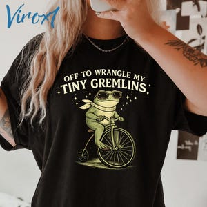 May include: Black t-shirt with the text "OFF TO WRANGLE MY TINY GREMLINS." The graphic features a frog wearing sunglasses and a scarf, riding a bicycle. The frog is green, and the bicycle is black and white.