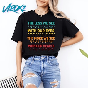 May include: Black t-shirt with the text "THE LESS WE SEE WITH OUR EYES THE MORE WE SEE WITH OUR HEARTS" in teal, yellow, orange, and red. The text includes braille dots. The shirt is worn with blue jeans.
