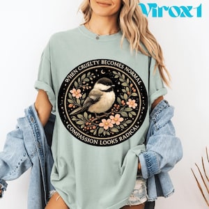 Puede incluir: Camiseta verde claro con un diseño circular. El diseño presenta una ilustración de un pájaro, flores y el texto "WHEN CRUELTY BECOMES NORMAL" y "COMPASSION LOOKS RADICAL".