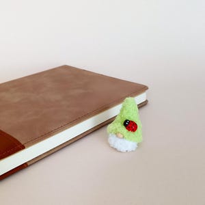 Può includere: Una piccola statuetta di gnomo realizzata con un tessuto verde chiaro soffice per il cappello a punta e lana bianca per la barba, dotata di un naso a perla di legno e una piccola coccinella rossa con punti neri applicata sul lato.