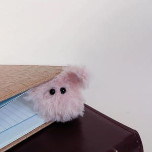 May include: A pink, fluffy creature bookmark with black eyes peeks out from between the pages of a brown leather-bound journal. The bookmark adds a whimsical touch to the book.