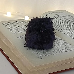 Pode incluir: Uma pequena criatura de pelúcia redonda de cor roxo-escura com pelo sintético longo e felpudo e dois olhos pequenos, escuros e brilhantes. A criatura tem uma aparência densa e texturizada e está posicionada nas páginas abertas de um livro.