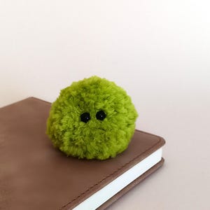 May include: A round, fuzzy, lime green pom-pom creature featuring two small, black, circular eyes positioned closely together on the front. The textured, yarn-based object has a soft, spherical appearance and is crafted from dense, vibrant green fibers.