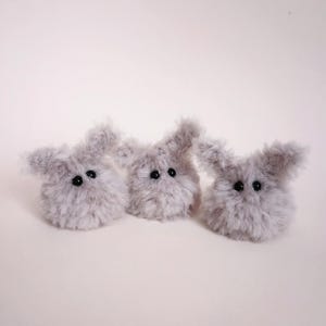 May include: Three fluffy, grey bunny-shaped plush toys with black button eyes are arranged on a light background. The plush toys have long, floppy ears and a soft, textured appearance. These handmade toys are perfect for Easter or as a gift.
