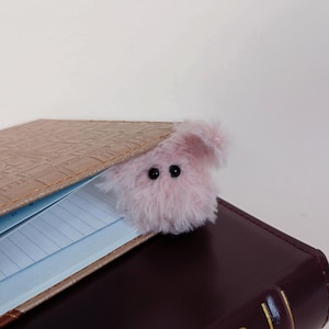 May include: A pink, fluffy creature with black eyes peeks out from between the pages of a brown leather-bound book. The book is open, revealing lined pages. The creature appears to be a bookmark or decorative item.