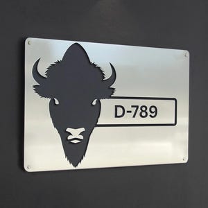 May include: A rectangular metal sign with a cut-out bison silhouette in black. The sign has the text "D-789" in a white rectangle. The sign is mounted on a dark gray wall.