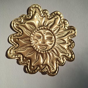 Puede incluir: Decoración de pared dorada en forma de sol con una cara sonriente, ojos cerrados y un detalle en forma de corazón. El sol tiene rayos detallados y un borde festoneado. El metal tiene un acabado pulido, reflejando la luz.