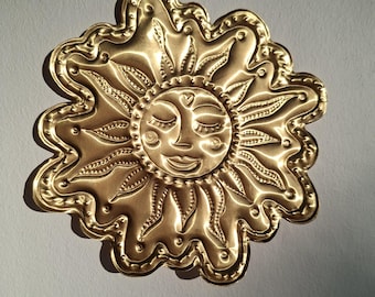 Puede incluir: Decoración de pared dorada en forma de sol con una cara sonriente, ojos cerrados y un detalle en forma de corazón. El sol tiene rayos detallados y un borde festoneado. El metal tiene un acabado pulido, reflejando la luz.