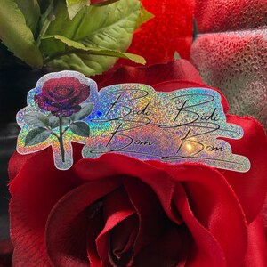 May include: A holographic sticker with a rose design and the words "Bidi Bidi Bom Bom". The sticker is set against a backdrop of a red rose and green leaves.