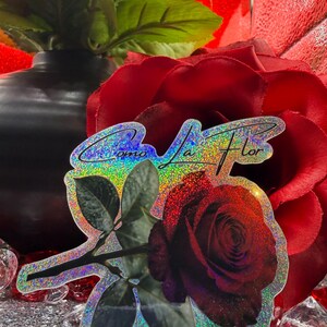 Op de afbeelding: Een holografische sticker met een donkerrode roos met groene bladeren en de tekst "Como la Flor". De sticker heeft een glinsterende, iriserende afwerking en staat tegen een achtergrond van rode rozen en een zwarte vaas.