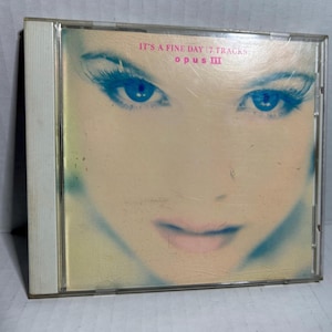 May include: A CD case featuring a close-up of a woman's face with striking blue eyes and long eyelashes. The text "IT'S A FINE DAY 7 TRACKS" and "opus III" are visible on the cover. The overall color scheme is soft and muted.
