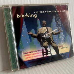 Puede incluir: Un CD de música B.B. King Let The Good Times Roll en una caja de plástico transparente. La portada muestra a B.B. King sentado en un taburete tocando una guitarra eléctrica negra frente a un horizonte urbano con estrellas blancas.
